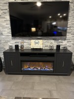 Mounted entertainment setup with TV on the wall above a dark gray fireplace console; decorative items and lighting in a showroom setting.